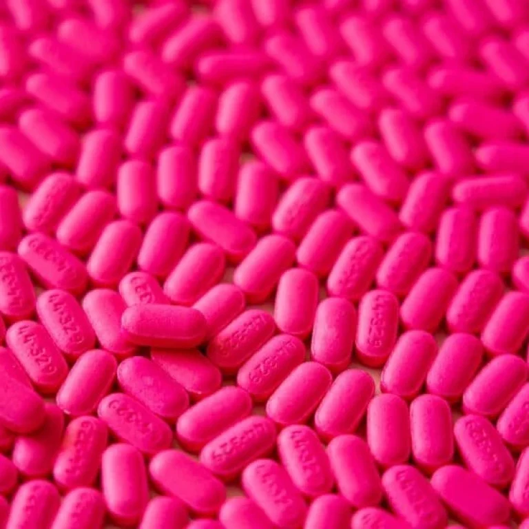 A large number of bright pink, oval-shaped tablets—possibly supplements such as Vitamin B12 vs B Complex—are spread out across a surface.