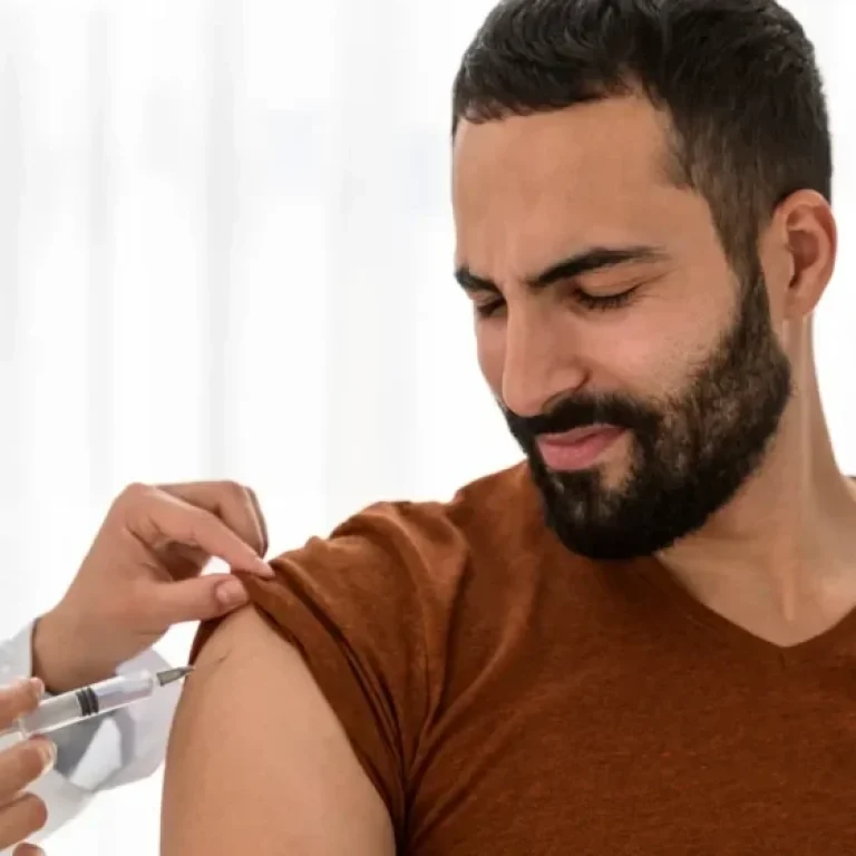 A person in a brown shirt receives a Vitamin D injection in the upper arm from a healthcare professional.