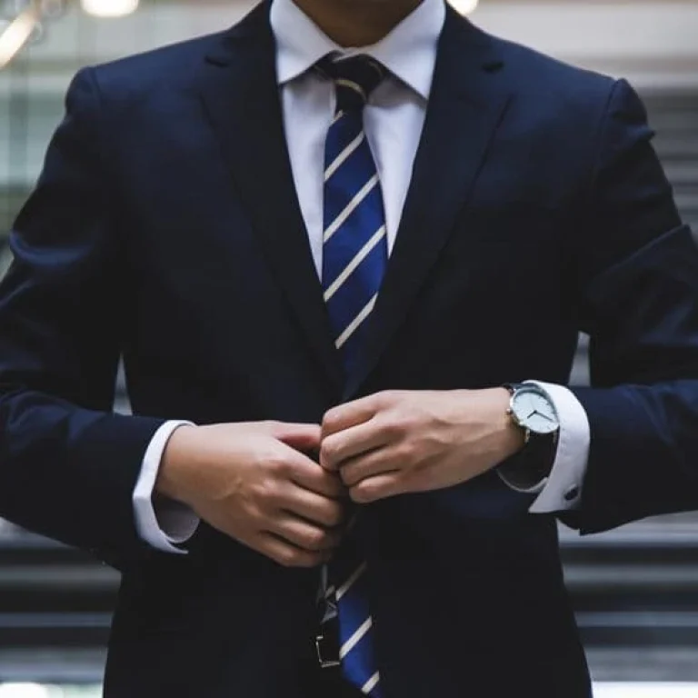 Person in a dark suit and striped tie fastens their jacket while standing in front of a staircase, exuding a professional edge.