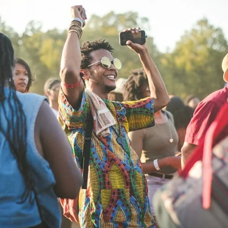 A person wearing colourful clothing and sunglasses raises their hands whilst dancing in a crowd at a summer festival. Trees are visible in the background, capturing the excitement of festival season.