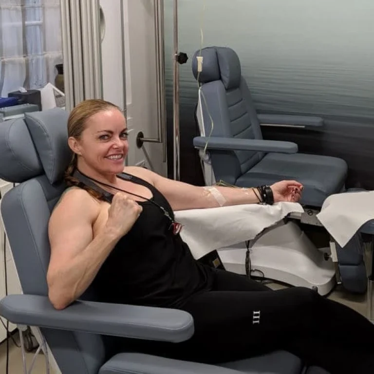 A woman sits in a medical chair at IV Boost Clinic, donating blood or plasma, smiling and flexing her arm with a drip line attached.