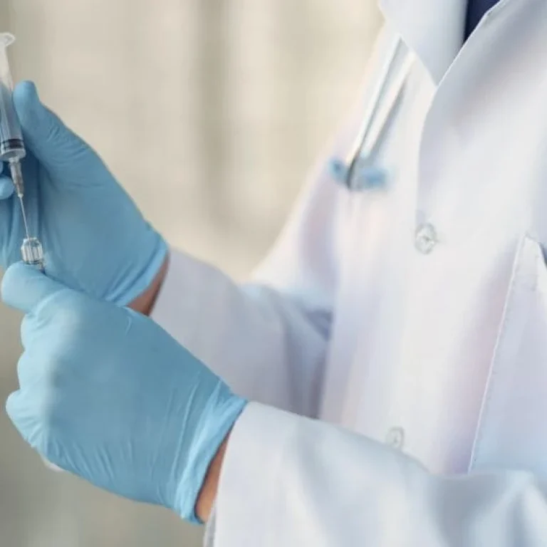 A healthcare professional wearing a white coat and blue gloves prepares a syringe with liquid Vitamin B12, with a stethoscope draped around their neck.