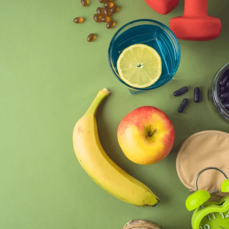 Banana, apple, a glass of water with lemon, dumbbells, supplements, headphones, sleep mask, and alarm clock arranged on a green surface—a perfect snapshot of biohacking essentials for a healthier lifestyle.