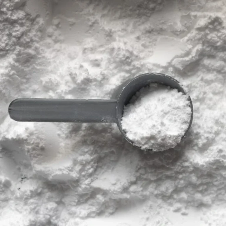 A grey plastic scoop filled with white powder, likely a glutamine supplement, rests on a surface covered in the same powder.