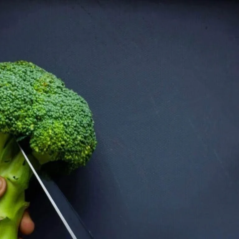 A hand holds a head of broccoli while a knife slices through the stem against a dark background—a nod to healthy eating and natural sources of antioxidants like glutathione.