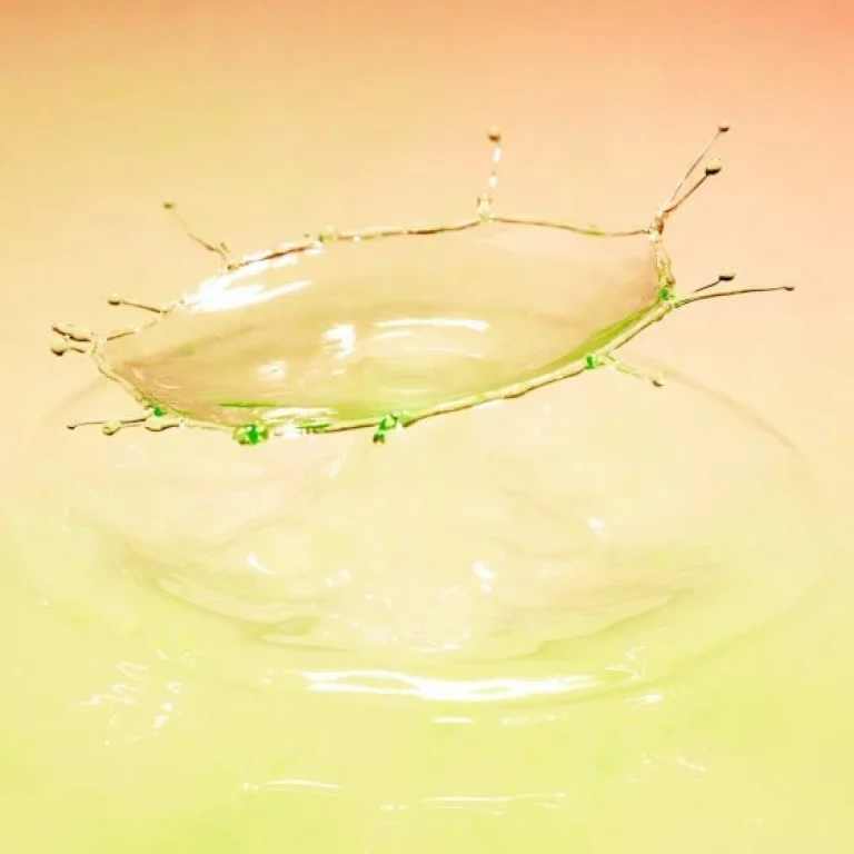 A high-speed photo captures a water droplet splashing, forming a crown-like shape against a soft pink and green gradient background—evoking the playful colours of a banana bag.