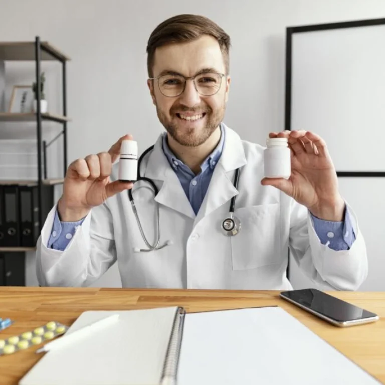 A male doctor wearing a white coat and stethoscope sits at a desk, smiling and holding two medicine bottles—one labelled vitamin B12 injection—with a notebook, mobile, and tablets on the table.