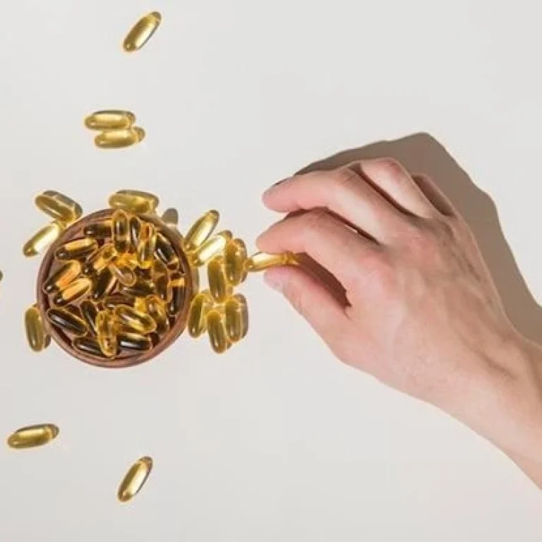A hand reaching for a yellow Vitamin B12 capsule from a small bowl filled with similar capsules, highlighting the importance of health, with some capsules scattered on a white surface.