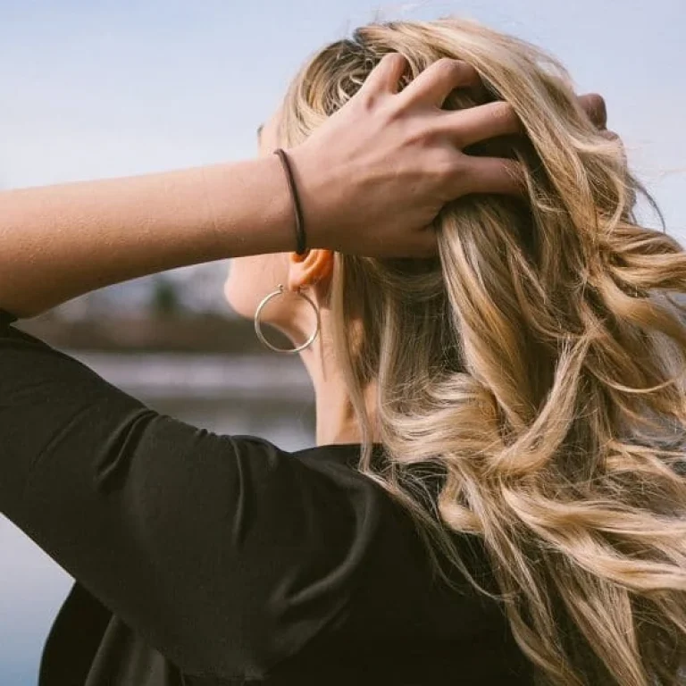 A person with long, wavy blonde hair stands outdoors near water, facing away and running a hand through their hair—a gentle reminder that prioritising hair care could be the perfect New Year’s Resolution. They wear a black top and a hoop earring.