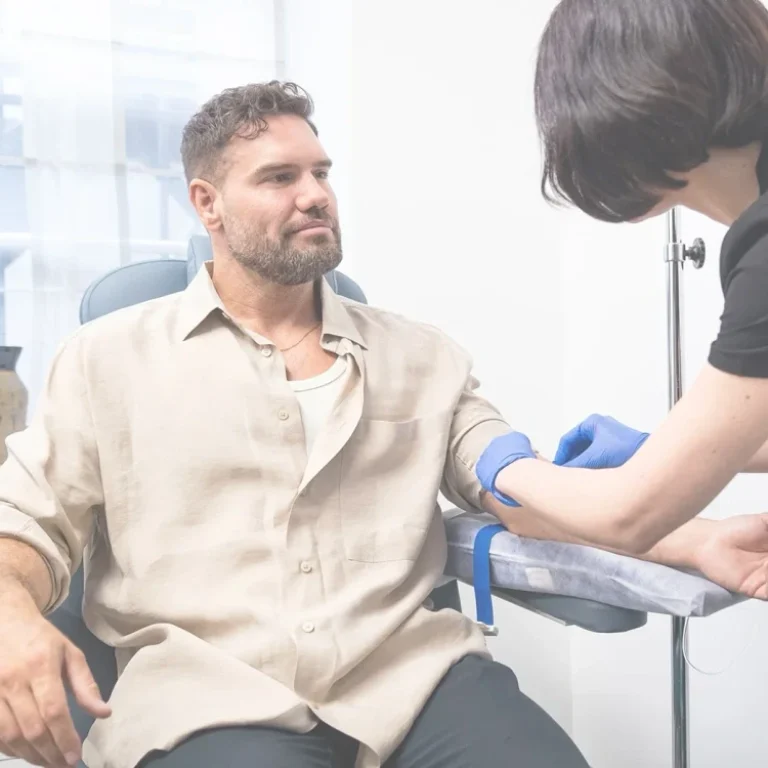 A man sits while a healthcare worker prepares to take blood from his arm in a clinical setting, where Anti-Ageing IV Booster treatments are also offered.