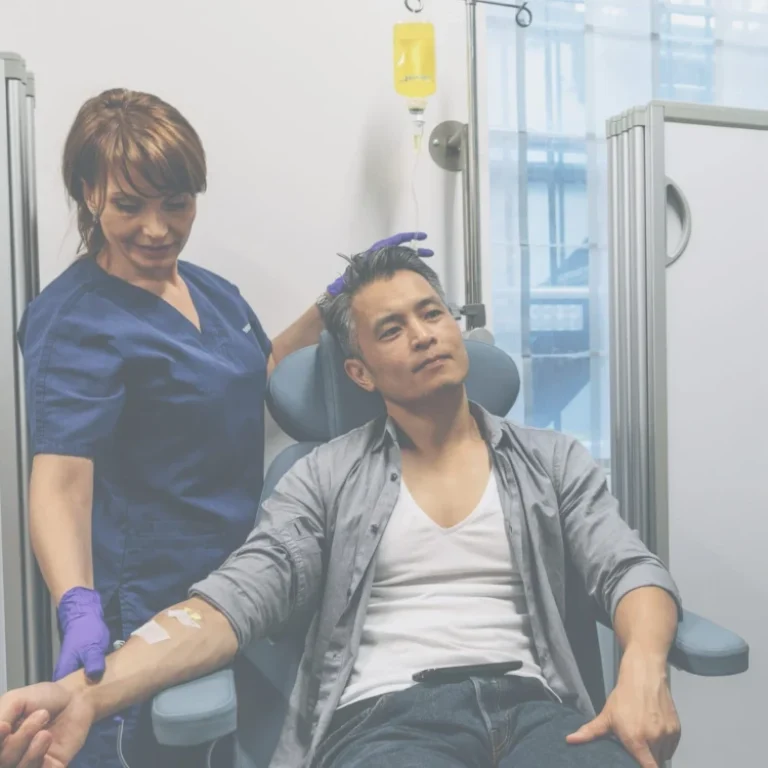 A nurse in blue scrubs assists a man receiving an IV therapy drip whilst seated in a medical clinic.