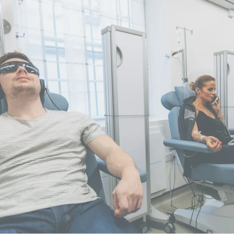 Three people sit in reclining chairs receiving IV Therapy; one man wears headphones and dark glasses, a woman uses a laptop, whilst another man relaxes in the background, all experiencing an Energy IV Booster for a revitalising energy boost.