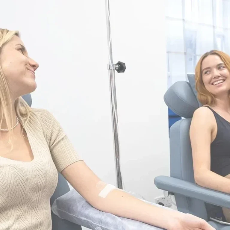 Two women sit in medical chairs, smiling at each other, with bandages on their arms in a clinical setting after receiving Hydration IV therapy.
